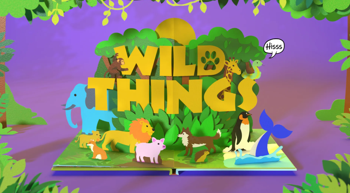 A colourful pop‑up book illustration with the words ‘Wild Things’ emerging from jungle foliage, surrounded by animals including an elephant, lion, giraffe, penguin, pig, fox, hedgehog and a whale, set against a purple background.