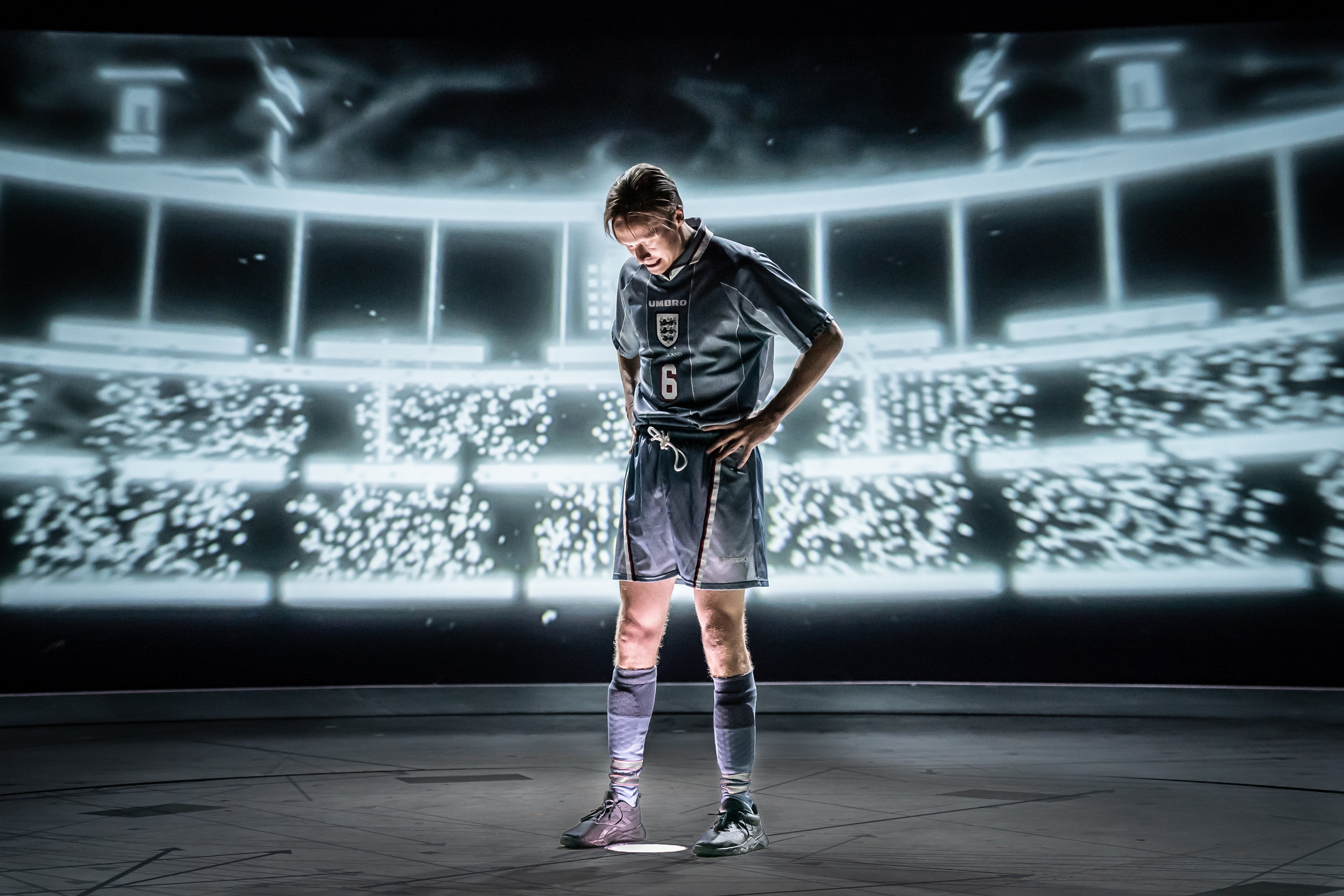 A promotional photo featuring an actor cast as Gareth Southgate dressed as a football player in an England kit with the number 6 on the jersey. He stands with hands on hips, looking down at the ground under a spotlight. The background is a large, black-and-white projection of a stadium filled with bright, blurred lights, creating a dramatic atmosphere. The stage floor is dark, emphasising the contrast between the performer and the illuminated backdrop.