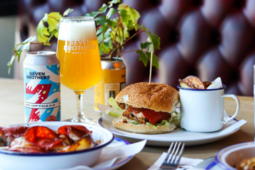An image of a burger with a Seven Bro7hers beer can and glass of beer in the background