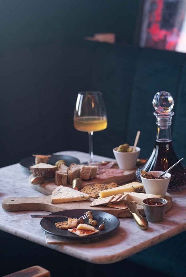 Photo of charcuterie of cheeses and meat, chutneys and olives, glass and carafe of wine laid out on a table.