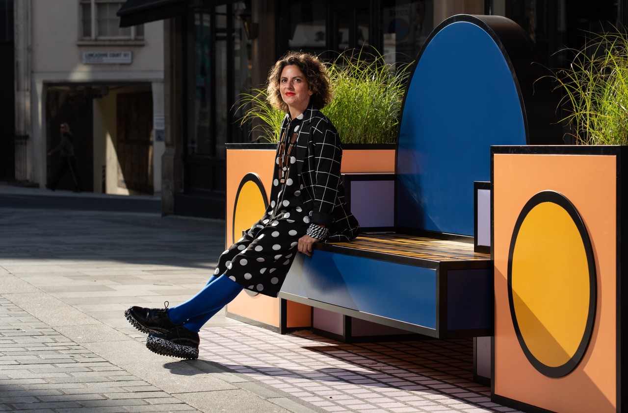 Photo of Camille Walala in a black and white polka dot outfit sitting on a colorful, modern bench adorned with vibrant blues, orange, and yellow geometric designs and greenery. The sun is shining.