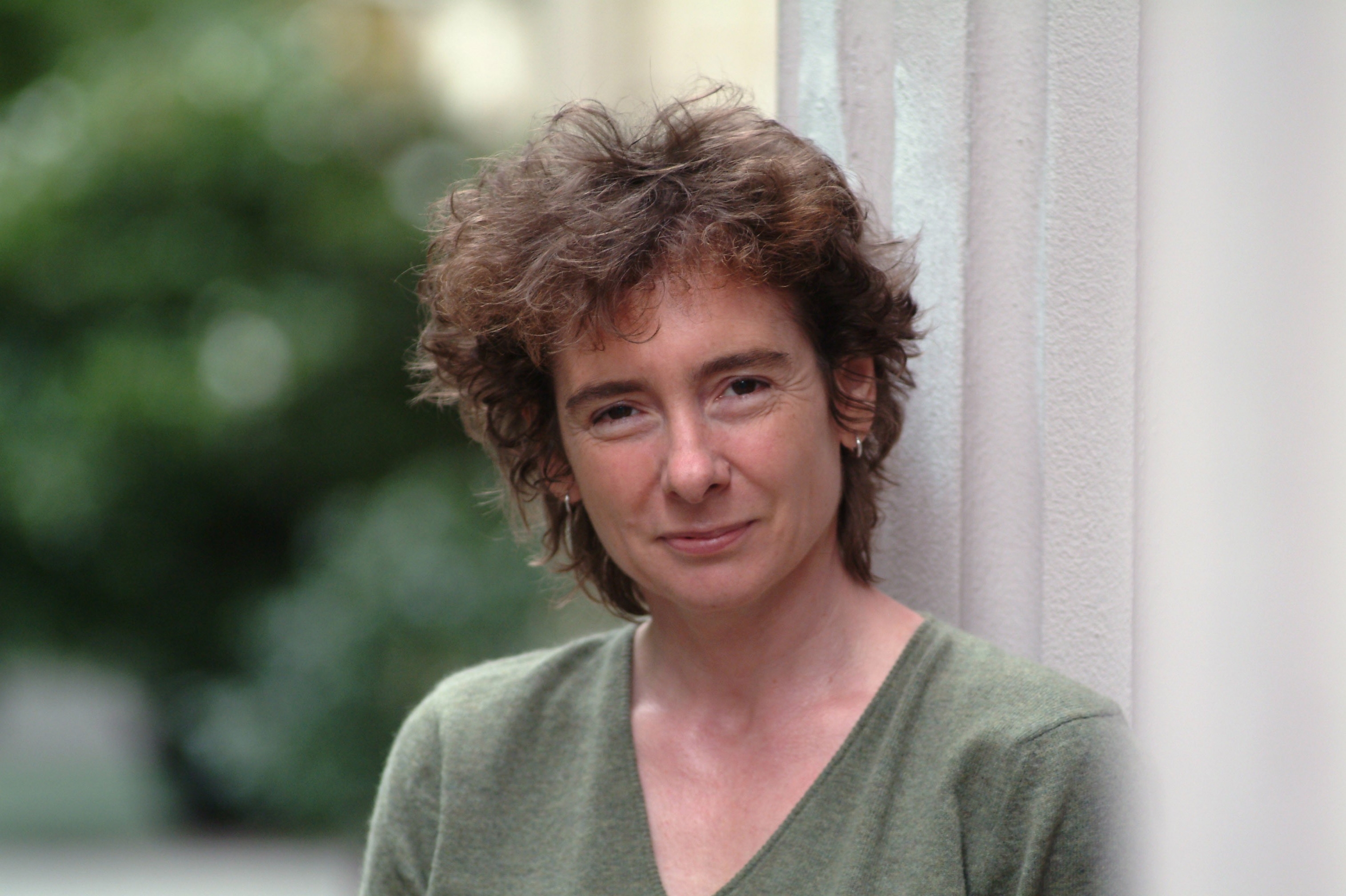 A photo portrait of Jeanette Winterson wearing a green shirt, leaning against a light gray wall, with a blurred background of greenery.
