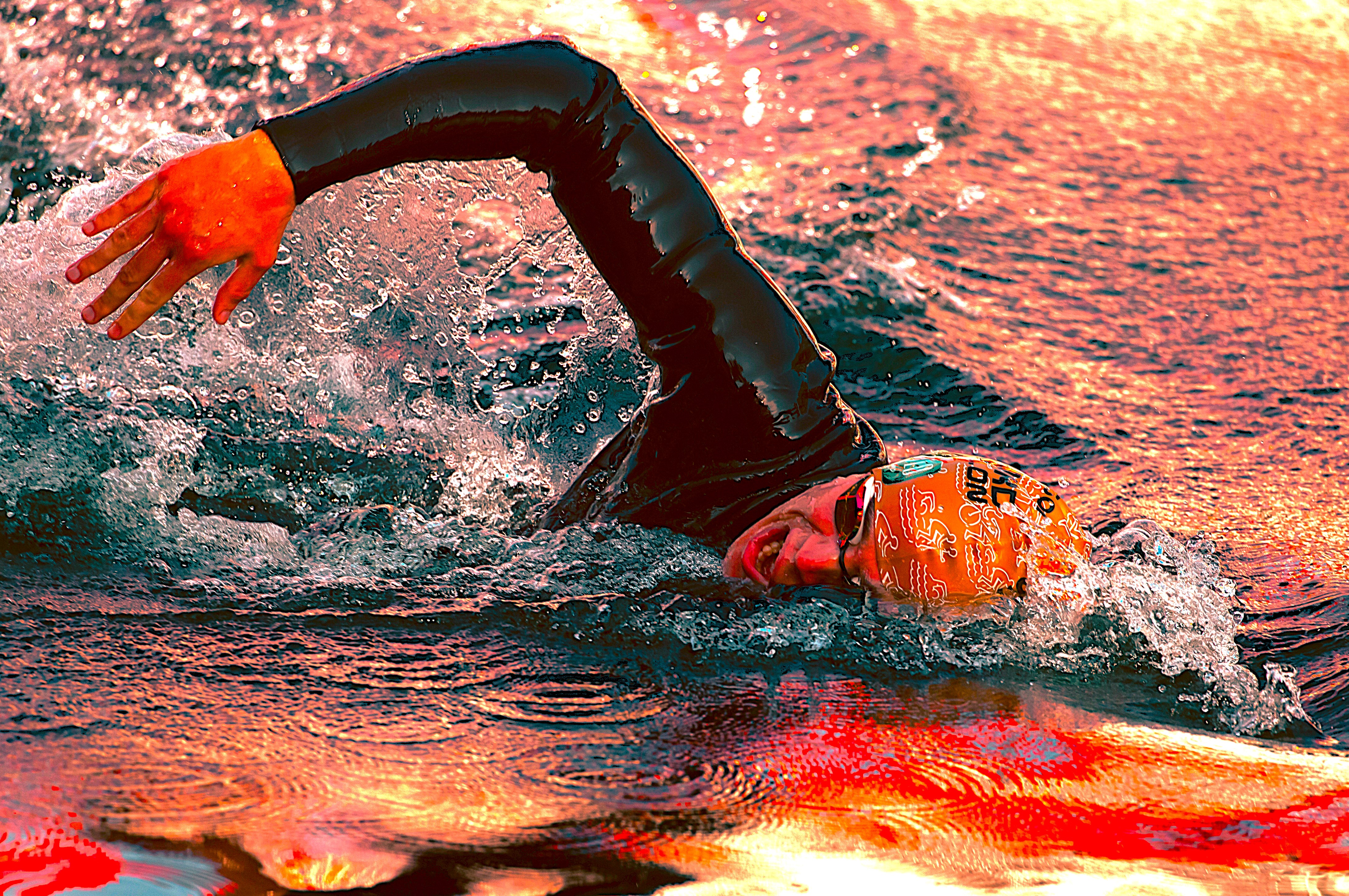 A swimmer wearing a black wetsuit, an orange swim cap, and goggles is captured mid-stroke during an open water swim. The athlete's arm is extended above the water, creating a splash as they propel forward. The water reflects a warm, fiery sunset, casting vibrant red and orange colours across the surface. The swimmer's face is partially submerged, with a determined expression visible through the goggles.