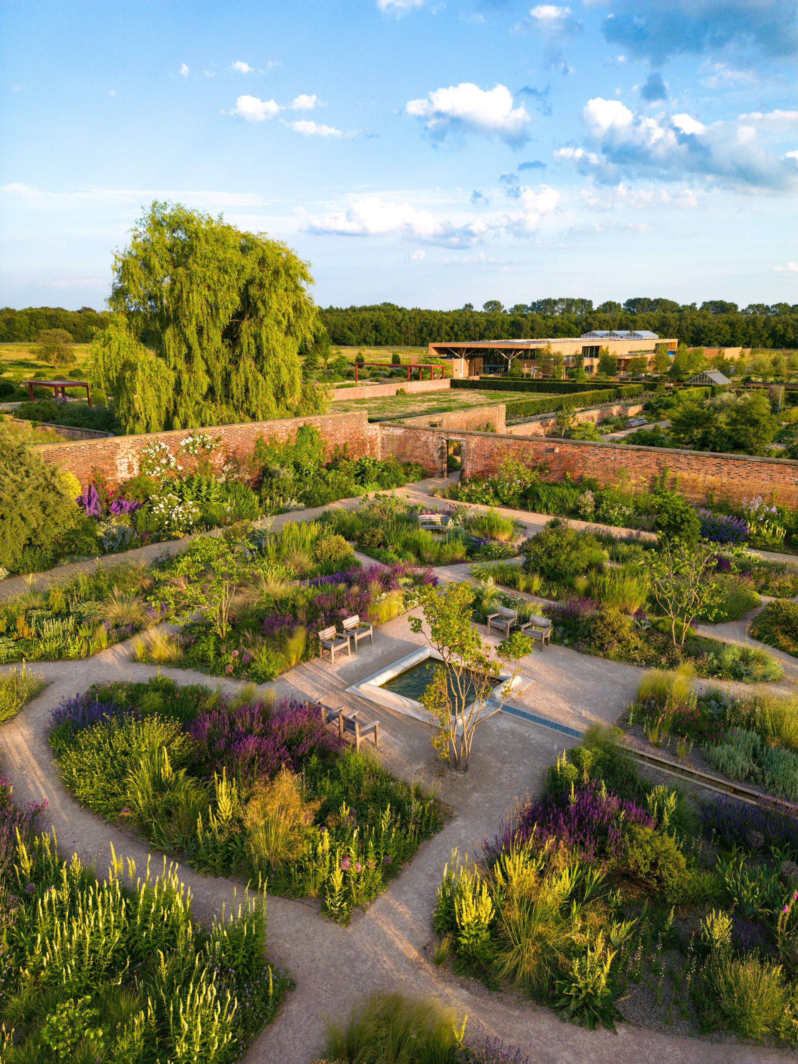 A formal garden with geometric paths, lush planting in green and purple tones, seating areas, and a central water feature, viewed from above on a sunny day.