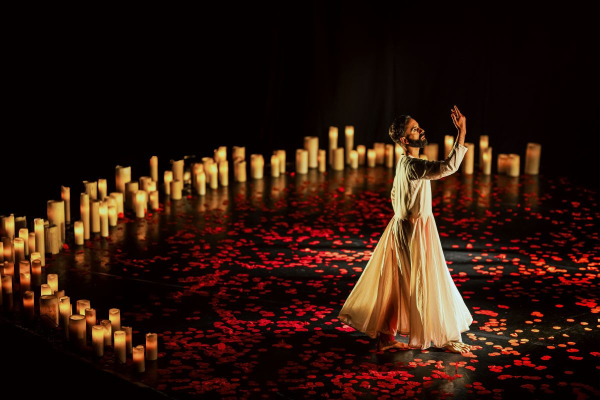 A male dancer in a flowing, cream-colored traditional outfit performs on a dimly lit stage. His arms are raised gracefully, and his expression is serene and focused. The stage is covered in scattered red rose petals, and a ring of glowing candles surrounds the performance space, casting a warm, golden light against the dark background.