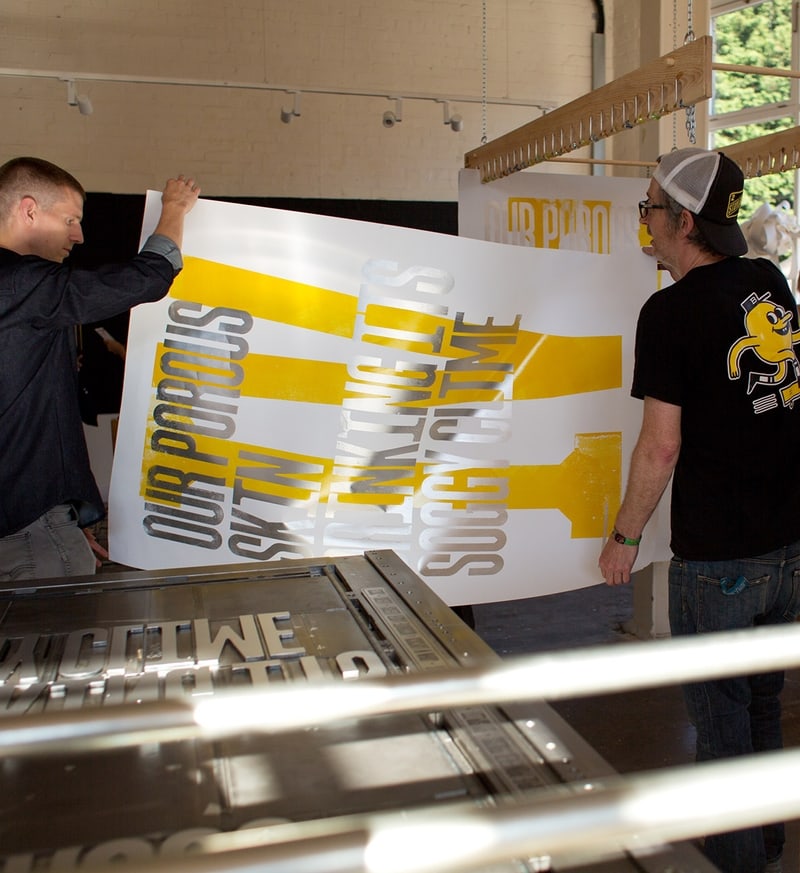 Two people holding a large poster with yellow and silver text that reads 'OUR POROUS SKIN THINKING IT'S SOCIETY TIME.' Their faces are blurred, and they are in a workshop or studio setting with various equipment and materials visible in the background.