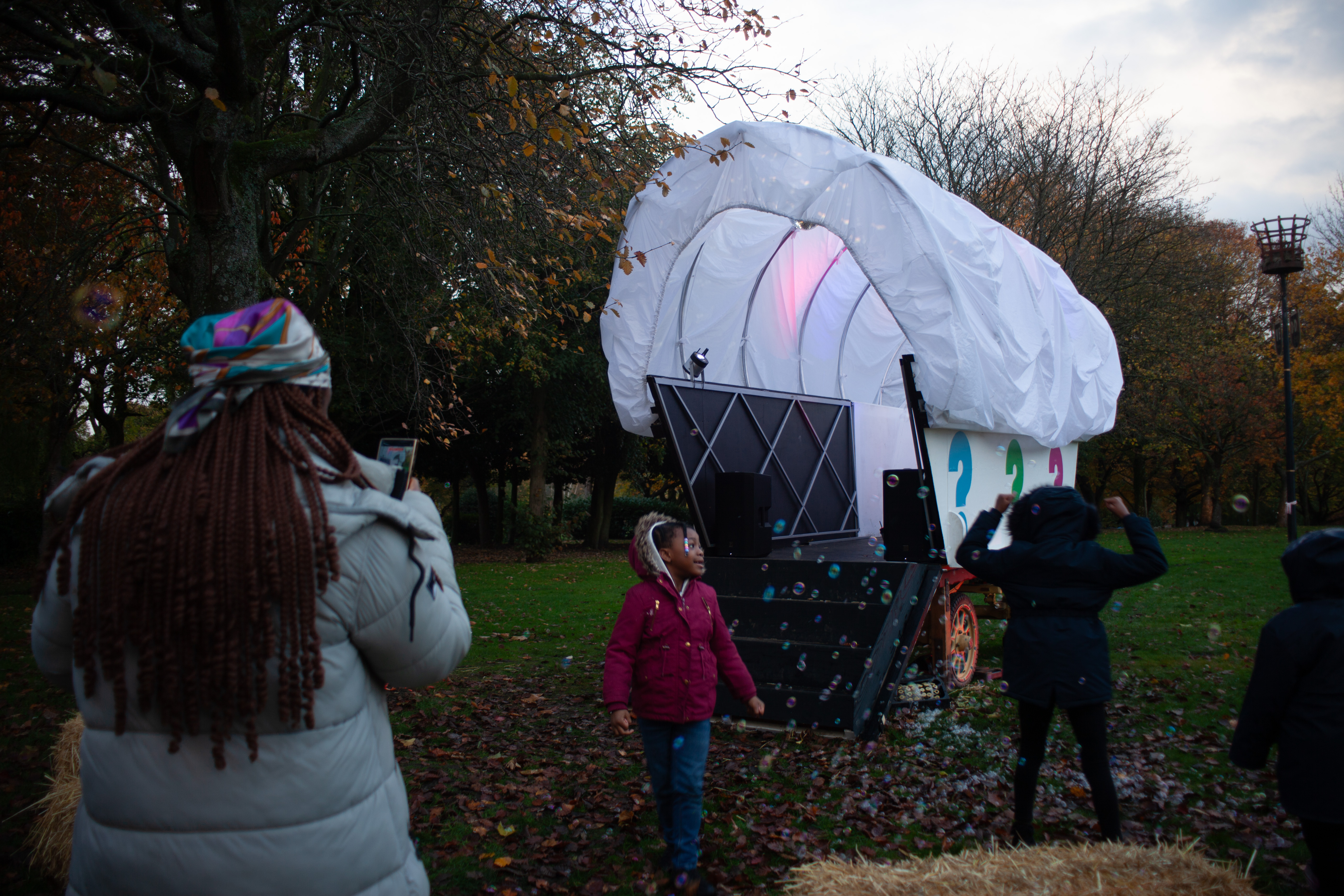 Outdoor scene in a park during autumn, showing a large white covered wagon-style structure with a rounded canopy and decorative panels. Several people stand nearby, including adults and children, with soap bubbles floating through the air. Trees with autumn leaves and grassy ground surround the installation