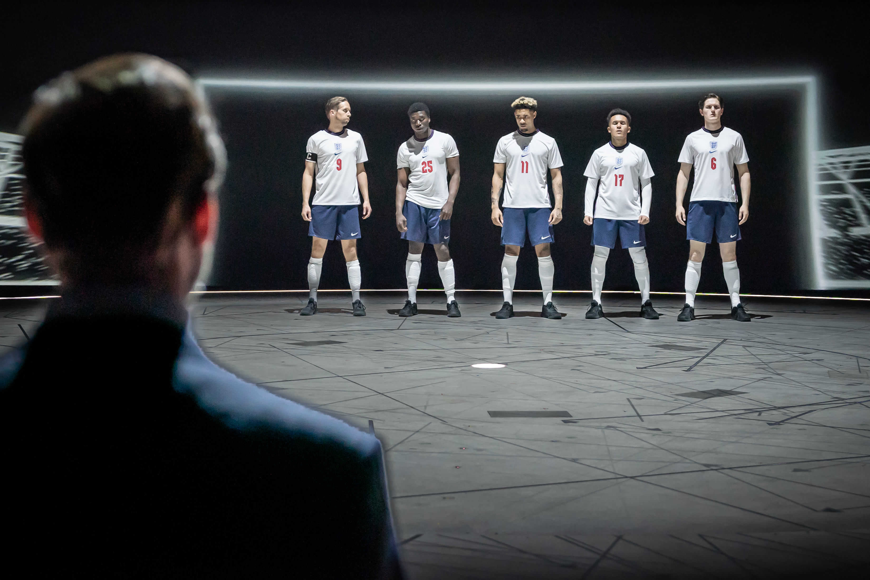 A promotional photo depicting the theatre production ‘Dear England’ – image shows five England football players in white jerseys stand in a dramatic spotlight, facing a suited individual, Gareth Southgate, in the foreground.