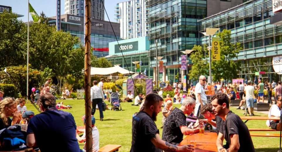 People enjoying a sunny day in a grassy urban plaza at MediaCity as part of We Invented the Weekend festival - surrounded by modern buildings, including those with BBC and dock10. Groups are sitting at picnic tables, walking, or lounging on the grass, with tents, stalls in the background.