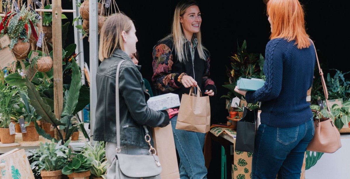 A group of three women are engaged in conversation at an outdoor market stall selling plants. The vendor, a woman with long blonde hair wearing a black floral-patterned jacket, is smiling while handing a brown paper bag to one of the shoppers. The stall is filled with potted plants and hanging kokedama (moss ball plants), creating a lush and inviting atmosphere.