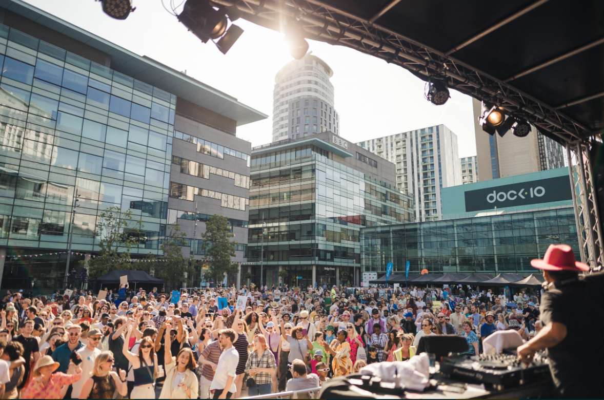 A large outdoor crowd gathered in front of a stage at MediaCity Piazza, surrounded by modern glass buildings. People are cheering and enjoying a sunny event while a DJ performs on stage in the foreground.