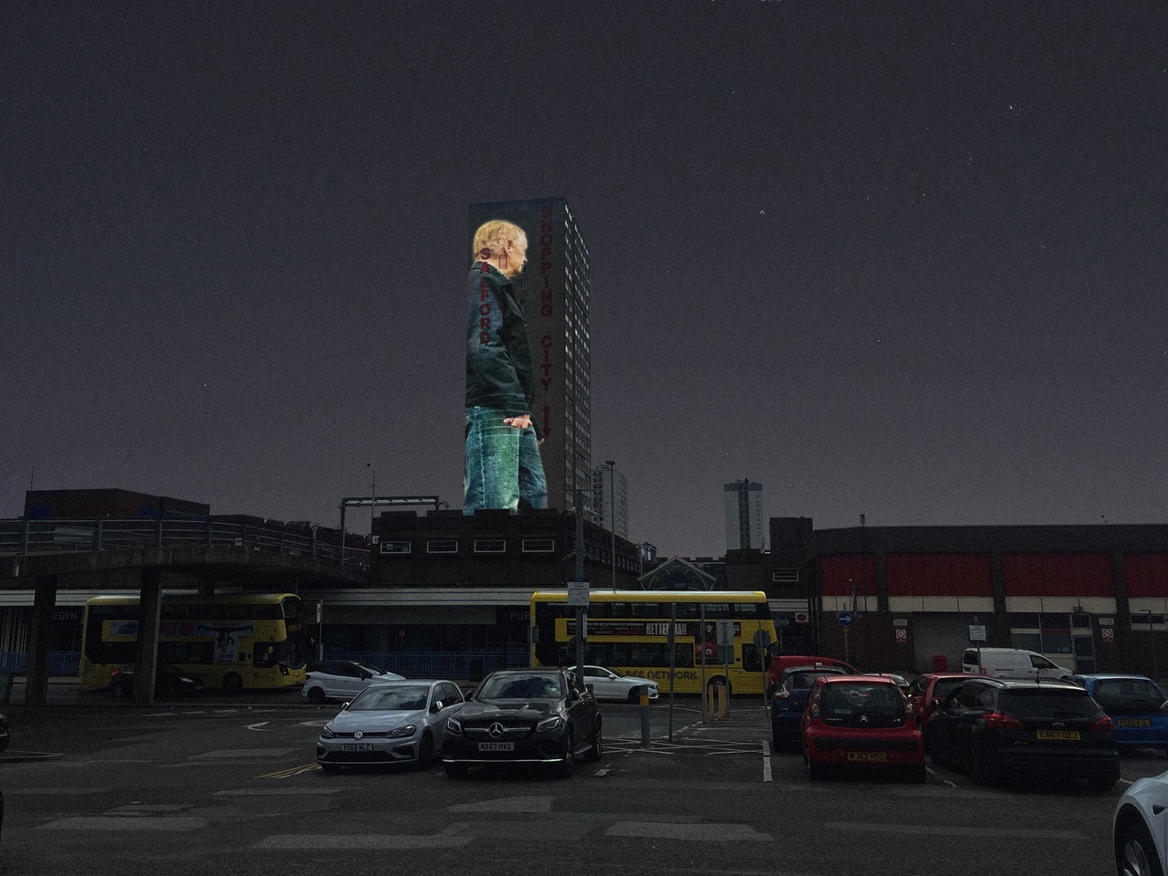 Night-time city scene with a tall tower block displaying a large projected image of a person in profile, with the words ‘Salford Shopping City’ running vertically down the building. In the foreground is a car park with parked cars and buses, surrounded by low-rise buildings under a dark, cloudy sky
