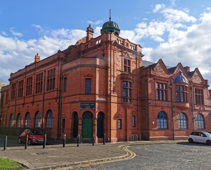 Historic redbrick building 'Salford Lads Club' with arched windows, a green door, and a small dome on the roof. The sun is shining, with a partly cloudy sky.