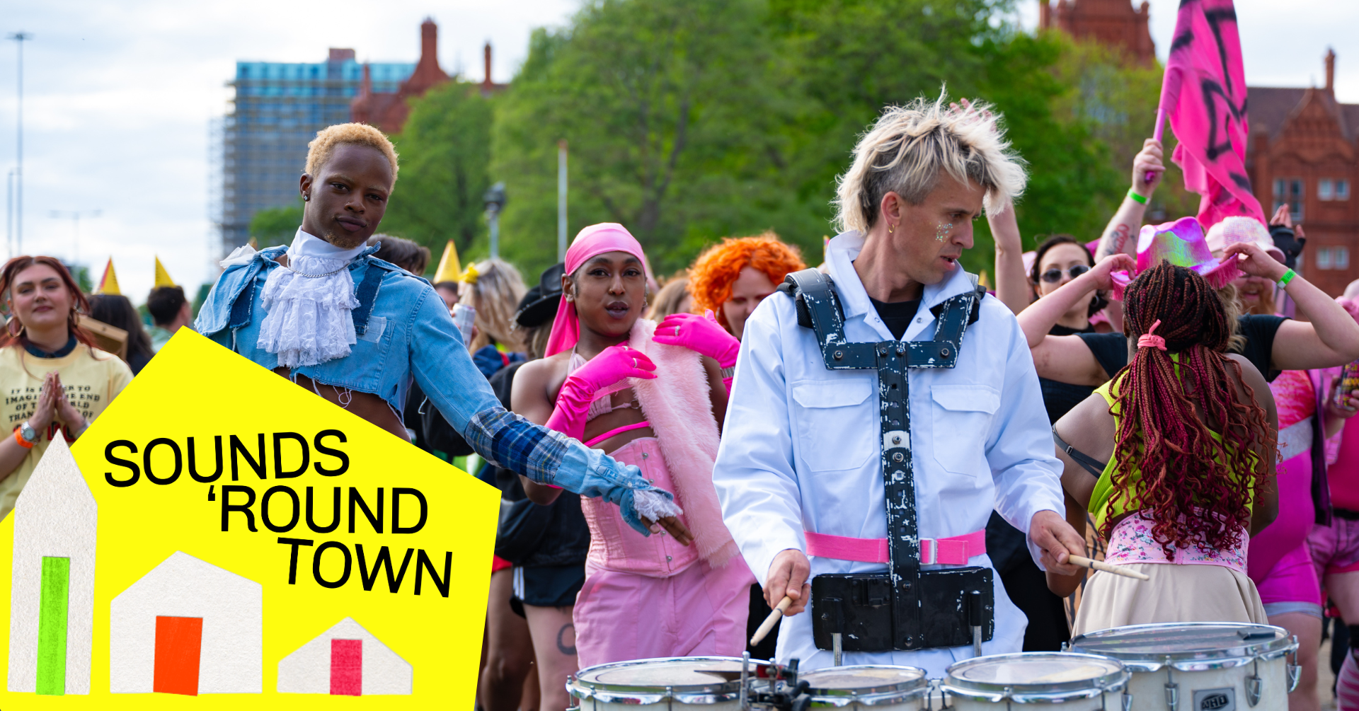 A lively outdoor parade scene featuring people in colourful and eclectic outfits. One person in a blue ruffled jacket and white lace cravat dances, while another in a white jumpsuit plays a marching drum. A third person in a pink corset and gloves sings or cheers. The crowd behind them is energetic, waving flags and dressed vibrantly. In the foreground, a bright yellow graphic with stylised houses reads “SOUNDS ‘ROUND TOWN.”