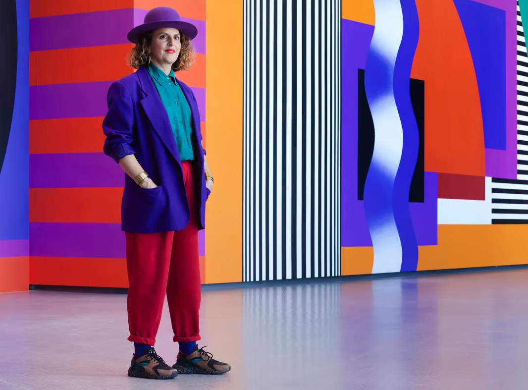 A person stands indoors against a vibrant, geometric mural featuring bold stripes, wavy shapes, and blocks of color in purple, orange, red, black, and white. The individual is dressed in a striking, colorful outfit: a purple wide-brimmed hat, a purple oversized blazer, a teal shirt, and bright red pants tucked into blue socks with dark sneakers.
