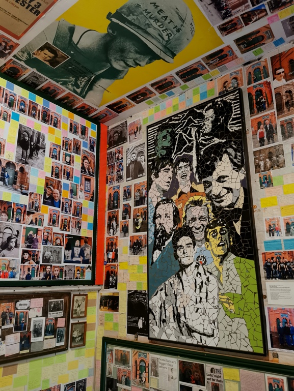 A room covered wall-to-ceiling with photos, notes, and memorabilia dedicated to The Smiths. A large mosaic portrait featuring famous musicians, including Noel and Liam Gallagher, is prominently displayed on one wall. The ceiling shows a large image of a soldier’s helmet with the words “MEAT IS MURDER.” Colorful sticky notes and framed photographs fill every available space, creating a dense, collage-like tribute. This is the Smiths Room at Salford Lads Club.