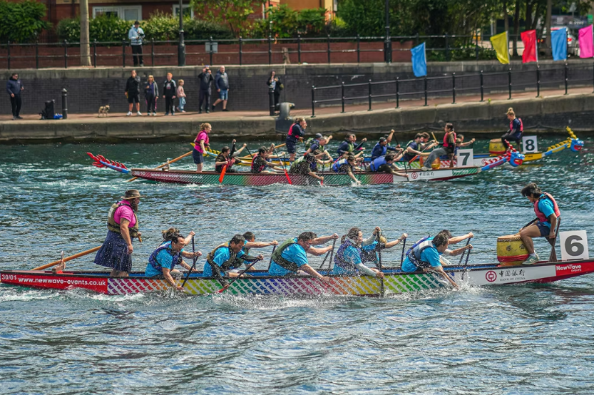 A dragon boat race at Salford Quays with three brightly decorated boats, each paddled by a team in matching shirts. The closest boat has a team in blue, paddling in sync while water splashes around them. Spectators stand at waterside, with colourful flags hanging nearby.