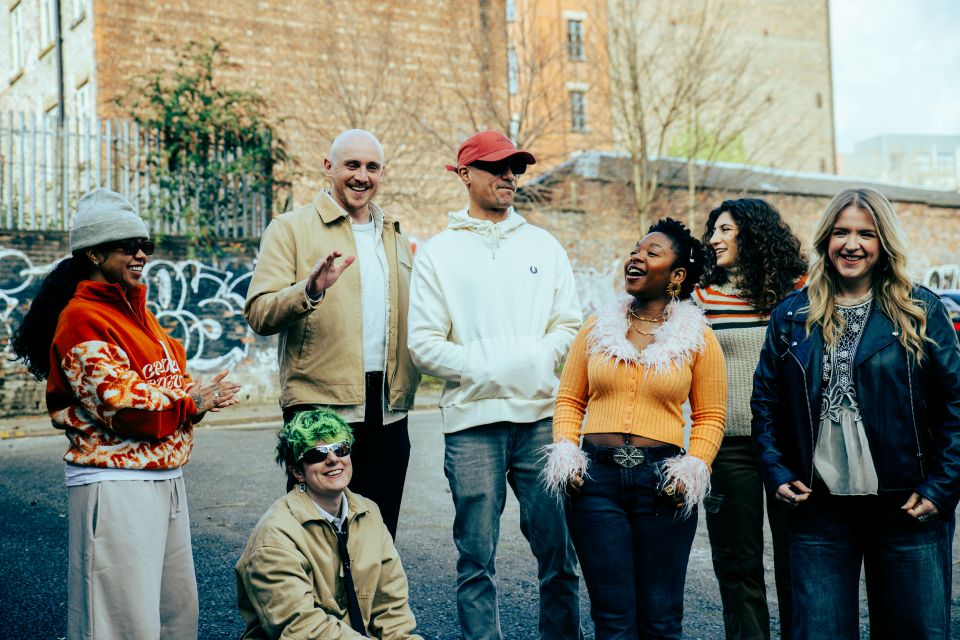 Group of people standing and crouching together on a street in an urban setting, with brick buildings and graffiti-covered walls in the background. The group wears a mix of casual and colourful clothing, including jackets, hoodies and knitwear. The scene appears informal and outdoors, photographed in daylight