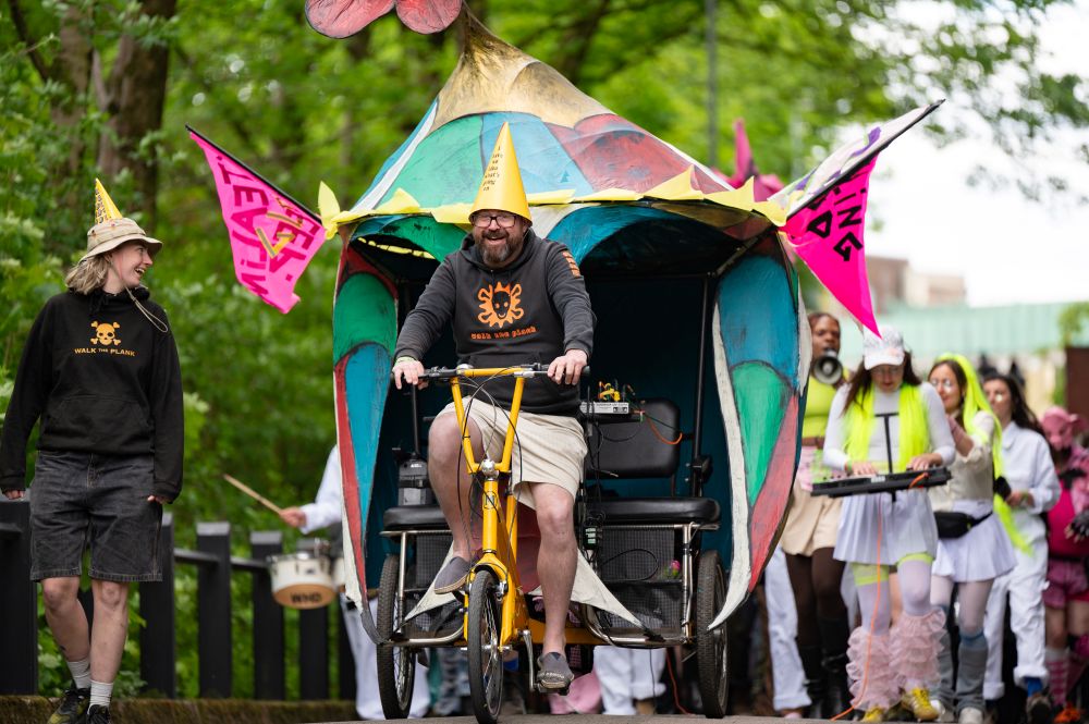 A joyful parade scene featuring a person riding a decorated tricycle with a large, colorful canopy structure. They wear a party hat and a “Walk the Plank” hoodie, smiling as they lead a group of performers. To the left, another person in similar attire walks alongside. Behind them, people dressed in bright costumes play instruments and wave pink flags among the trees.