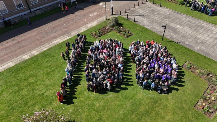 Aerial view of a large group of people standing on a green lawn arranged to form the number “100”, with surrounding paths, buildings and landscaped areas visible.