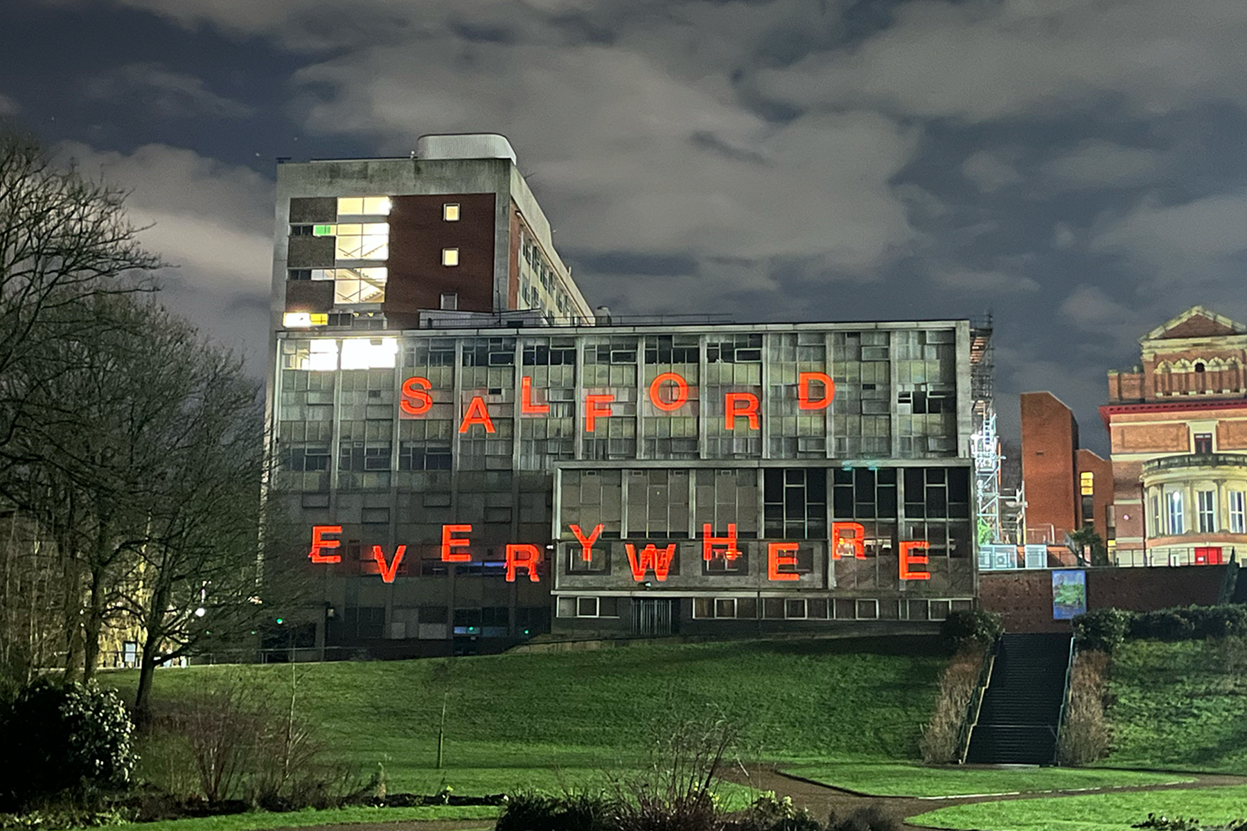 Night-time view of a large concrete building on a grassy slope, with bright red illuminated letters spelling ‘SALFORD EVERYWHERE’ across the façade. Trees, steps, and surrounding buildings are visible under a cloudy night sky.