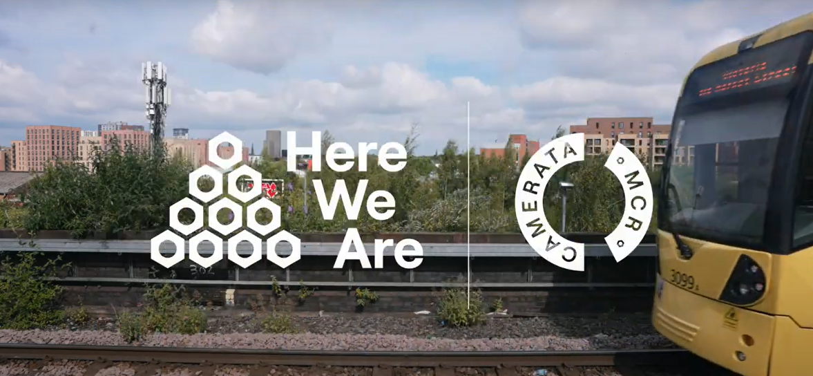A yellow tram on the right side with the white text 'Here We Are' and a hexagonal pattern on the left. A circular logo reading 'CAMERATA MCR' appears next to the text. The background features train tracks, greenery, and buildings beneath a partly cloudy sky.